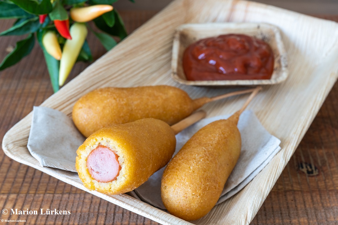 CORN DOG, Werbematerial Drei mit einem Teigmantel umhüllten Bockwürstchen auf einem Teller. Ein Corn Dog ist im Anschnitt zu sehen.